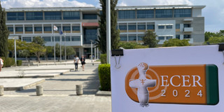 Foto des Universitätsgeländes mit einem Gebäude im Hintergrund und dem Plakat der ECER 2024 mit deren Logo im Vordergrund