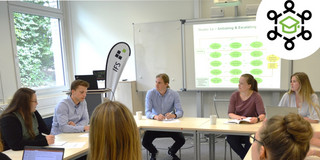 PromoKon Foto von mehreren Promovierenden an Seminartischen mit einer projezierten Präsentation am Whiteboard.