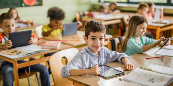 Grundschulkinder im Klassenraum, die mit Tablets und Schulheften arbeiten