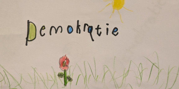 Demokratie Kinderzeichnung einer Wiee mit einer Blume Sonne und dem Schriftzug Demokratie